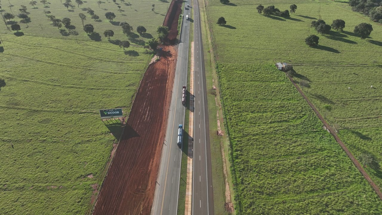 Vista aérea da BR-163 com terraplenagem para a nova pista ao lado da via existente, com caminhões em tráfego, em Mato Grosso do Sul