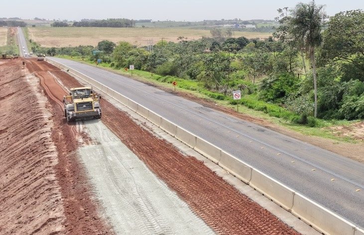 Nova faixa da BR-163 já com barreira de concreto instalada, com máquina compactando a base ao lado da pista existente