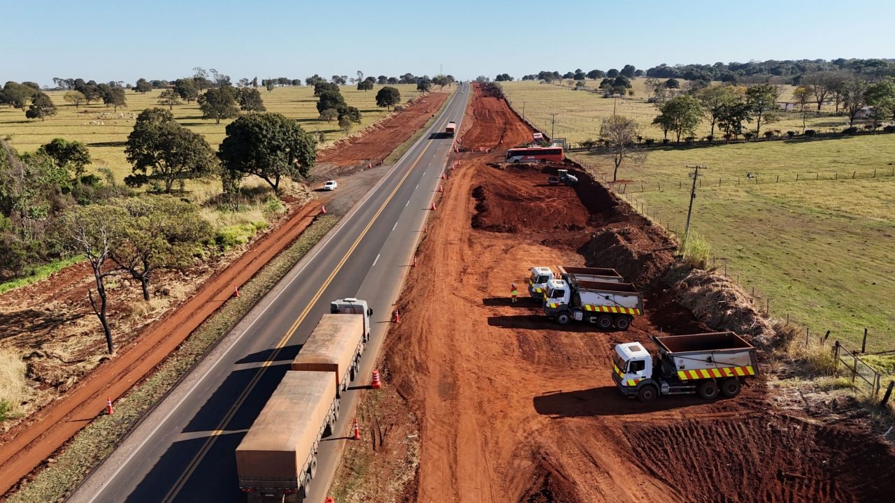 Vista aérea de obra de terraplenagem para a duplicação da BR-163, em Mato Grosso do Sul, com caminhões e tráfego de carga na pista existente